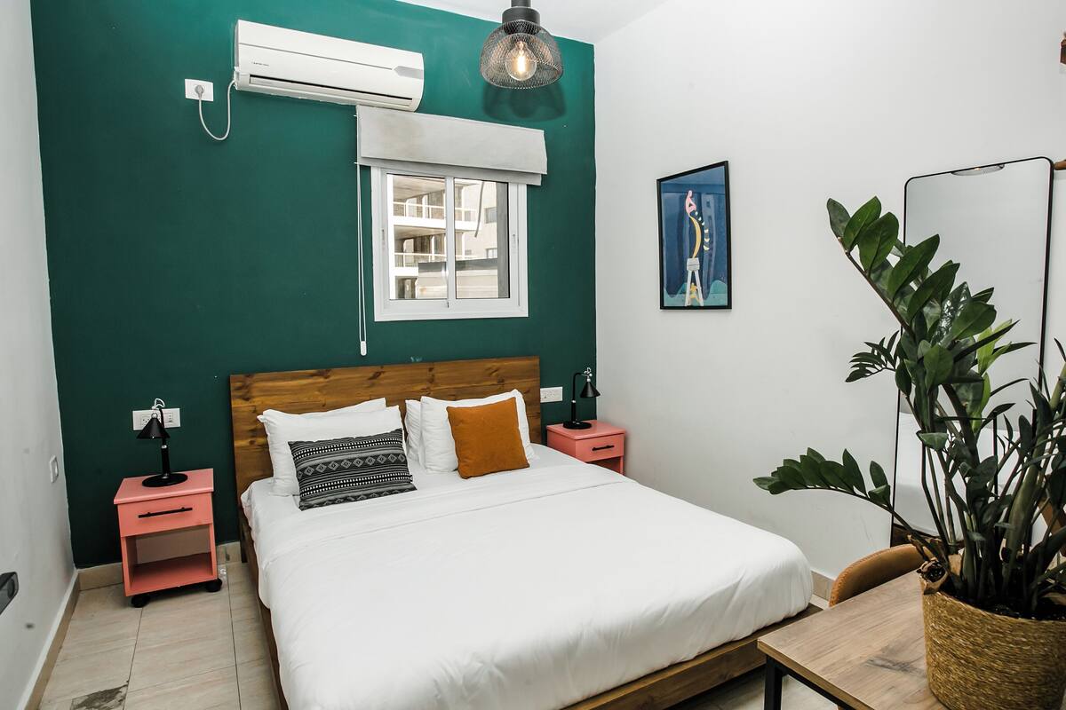 A bedroom with a bed, a nightstand, a bedside lamp, an air conditioner, a window, a wall-mounted picture, and a potted plant.