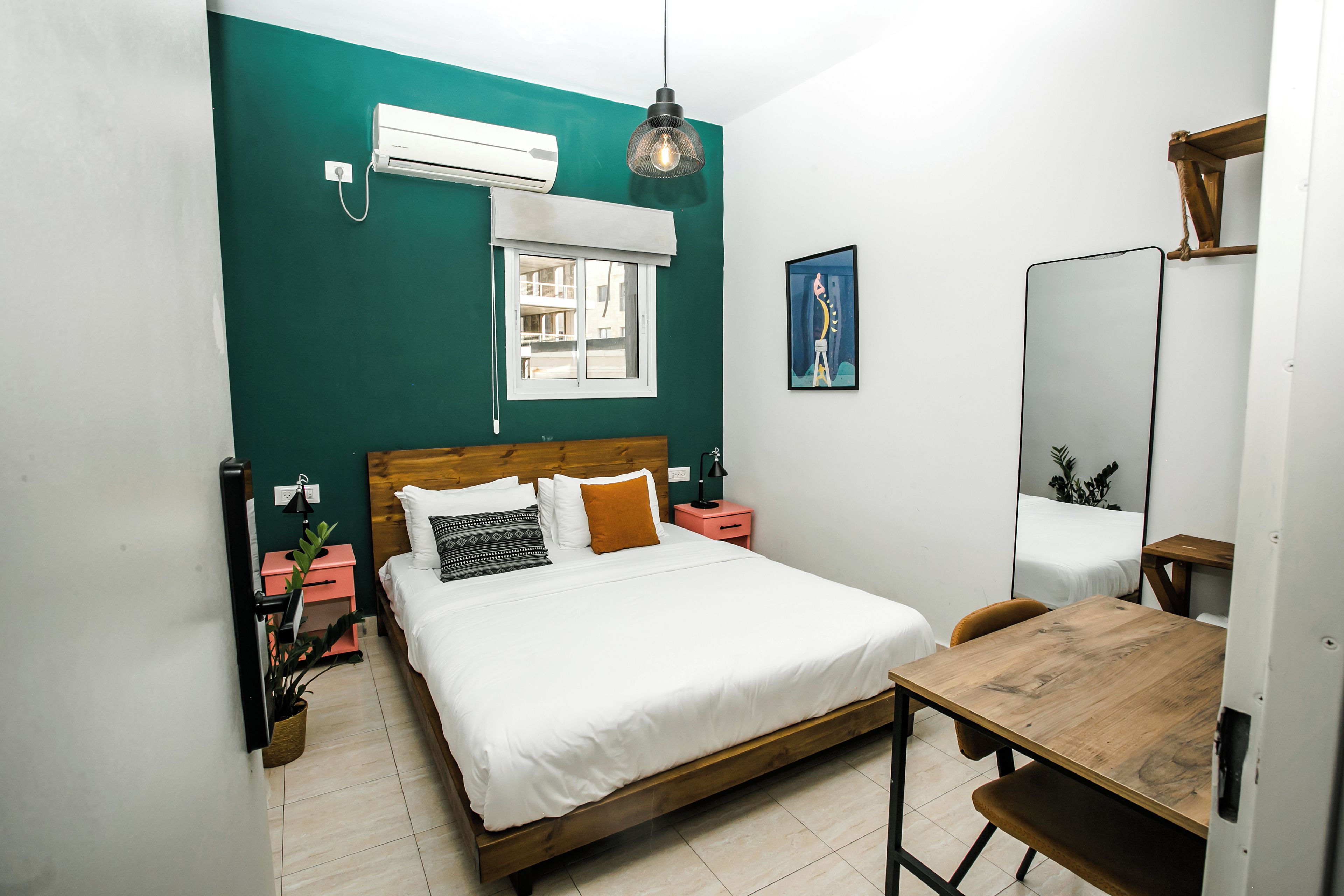 A bedroom with a bed, bedside tables, a desk, a chair, a hanging light fixture, a window, and an air conditioning unit.