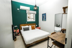 A bedroom with a bed, bedside tables, a desk, a chair, a hanging light fixture, a window, and an air conditioning unit. - Domus Tlv (Tel Aviv)