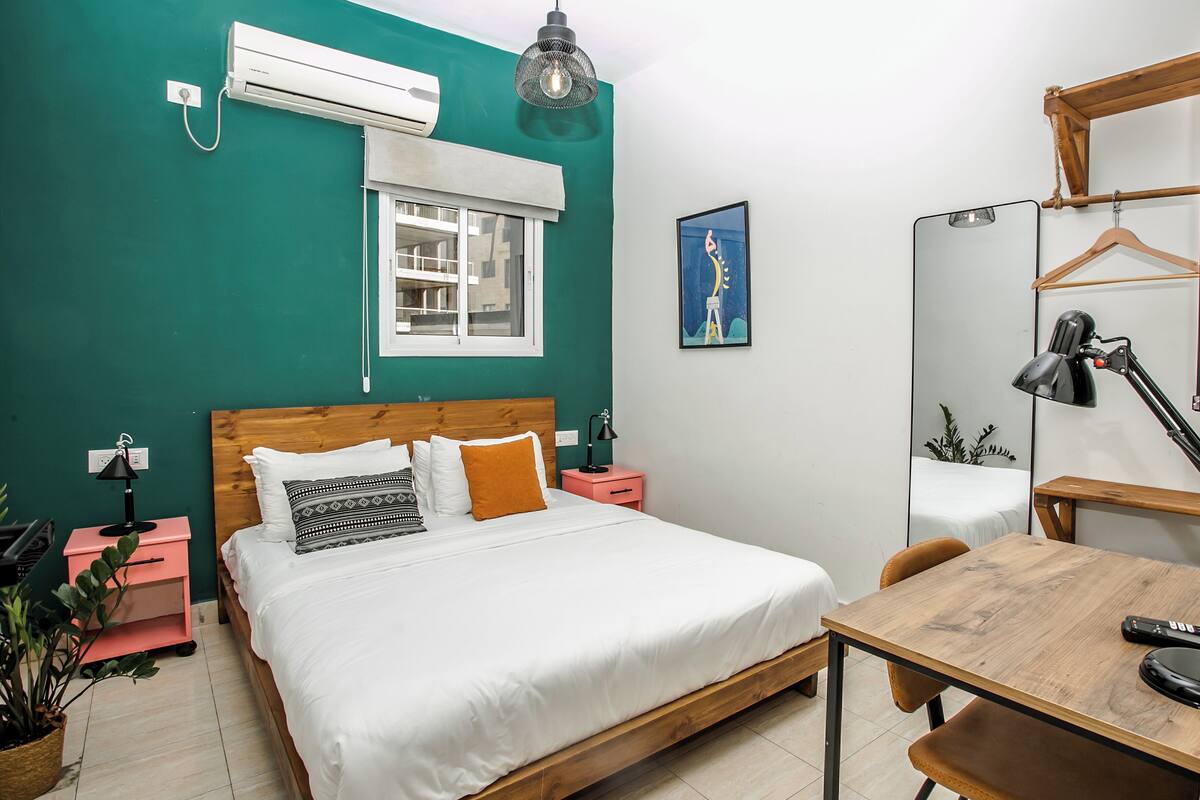 A bedroom with a bed, bedside tables, a desk, a mirror, and a window.