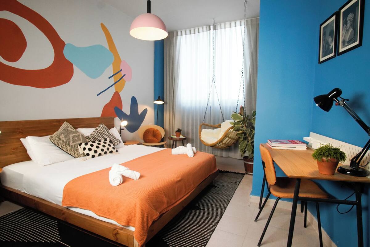 A modern hotel room with a large bed, a desk, a chair, a hanging chair, and a large abstract painting on the wall.
