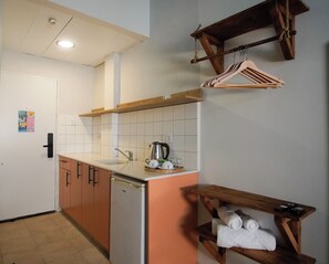 Une cuisine compacte équipée d’un évier, d’une machine à café et d’un petit réfrigérateur. On y trouve une étagère en bois avec des serviettes et un porte-serviettes suspendu au-dessus de l’évier.