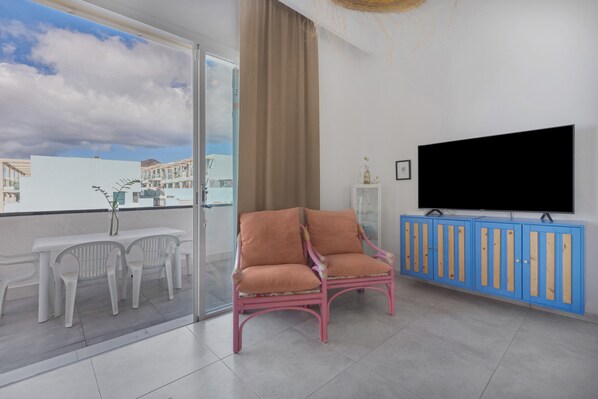 Een balkon met een witte tafel en stoelen, een roze stoel, een blauwe kast en een televisie aan de muur.