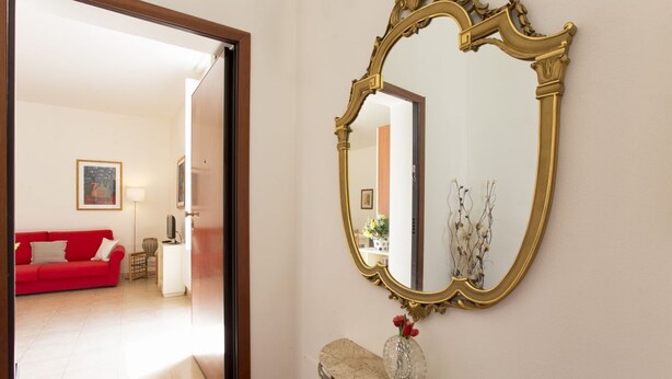 Um corredor com um sofá vermelho, uma mesa de apoio com um vaso e um espelho grande e ornamentado.