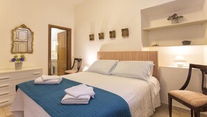 Quarto com cama, mesa de cabeceira, cadeira, espelho e prateleira com objetos decorativos.