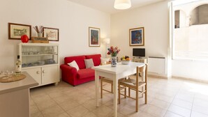 Uma sala de estar moderna com um sofá vermelho, uma mesa de jantar branca e uma televisão. Há quadros nas paredes e um vaso com flores sobre a mesa.