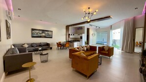 A modern living room with a sofa, coffee table, and armchairs, a dining area with chairs and a table, a kitchen, and a view of the outdoors. - Lamar Park Resort  (Riyadh)