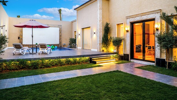 A modern outdoor pool area with lounge chairs and a red umbrella, surrounded by well-maintained landscaping and a building entrance with glass doors. - Lamar Park Resort  (Riyadh)