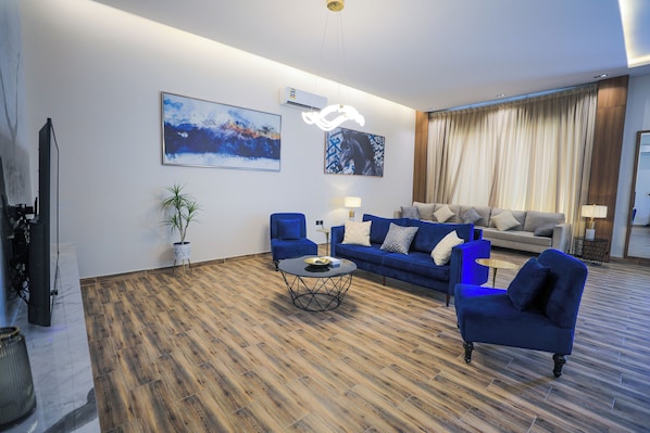 A modern living room with a wooden floor, a blue sofa, a blue armchair, and a coffee table. There are two wall-mounted air conditioners and a flat-screen TV on a stand. - Lamar Park Resort (Riyadh)