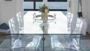 A modern dining area with a glass table and chairs, a potted plant, and a view of the sea through a large window. - Vashon Modern Waterfront Retreat+Private Beach+BBQ+Firepit (Vashon)