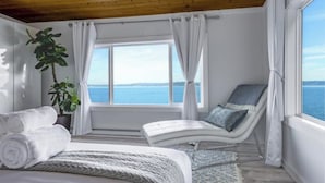 A bedroom with a curved bed, a reclining chair, a view of the sea, and a wooden ceiling. - Vashon Modern Waterfront Retreat+Private Beach+BBQ+Firepit (Vashon)
