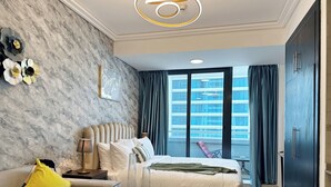 A modern hotel room with a marble wall, a large bed, a sofa, a glass table, and a view of a building through the window. - Gcv2 (Dubai)