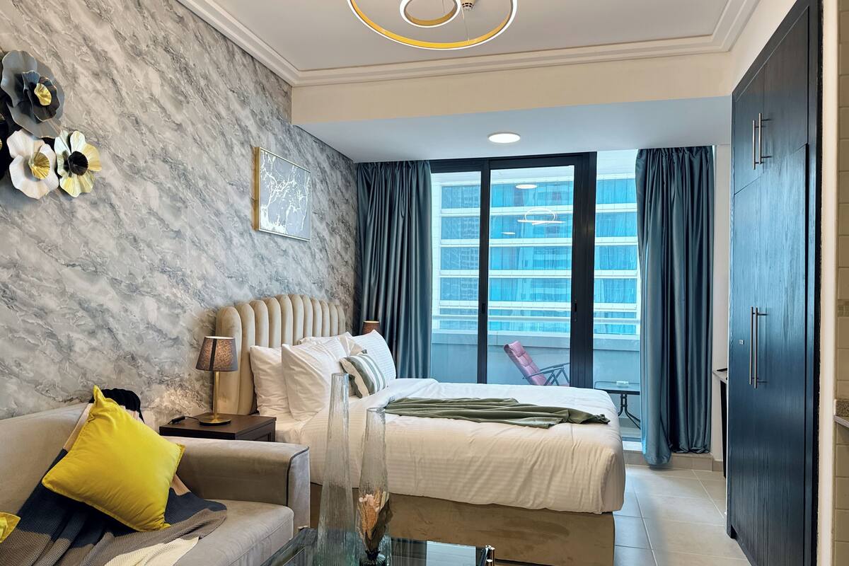 Une chambre d’hôtel moderne avec un mur en marbre, un grand lit, un canapé, une table en verre et une vue sur un immeuble par la fenêtre.