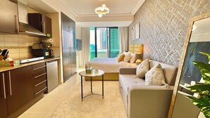 A compact hotel room with a kitchenette, a bed with a headboard, a sofa, a small round table, and a large mirror. - Gcv2 (Dubai)