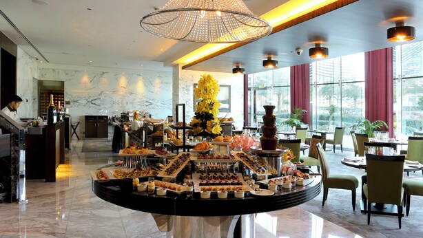 A hotel lobby with a marble floor, a central buffet with various food items, a chandelier, and large windows with curtains. - Gcv2 (Dubai)