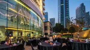 An outdoor dining area with round tables and chairs, set against a modern building with large windows. - Gcv2 (Dubai)
