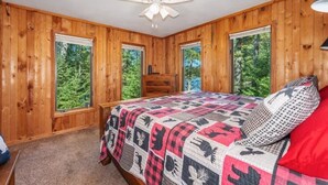 A bedroom with wooden walls, a bed with a quilt, a ceiling fan, and windows with a forest view. - Eagle River Clover Pines Holiday Home (Eagle River)