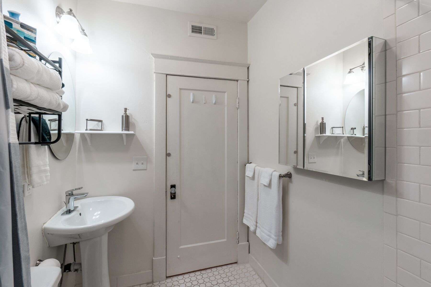 Une salle de bain avec un lavabo blanc, un miroir et une porte. Des serviettes sont accrochées au mur, et il y a un porte-serviettes.