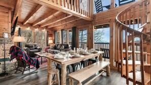 Interior de una cabaña de madera con una mesa de comedor para cuatro personas, una mecedora, un sofá y una escalera.