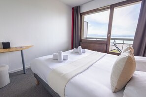 A hotel room with a bed, a wooden side table, a window with a view, and a red curtain. - Côté Lac Hostel (Neuvecelle)