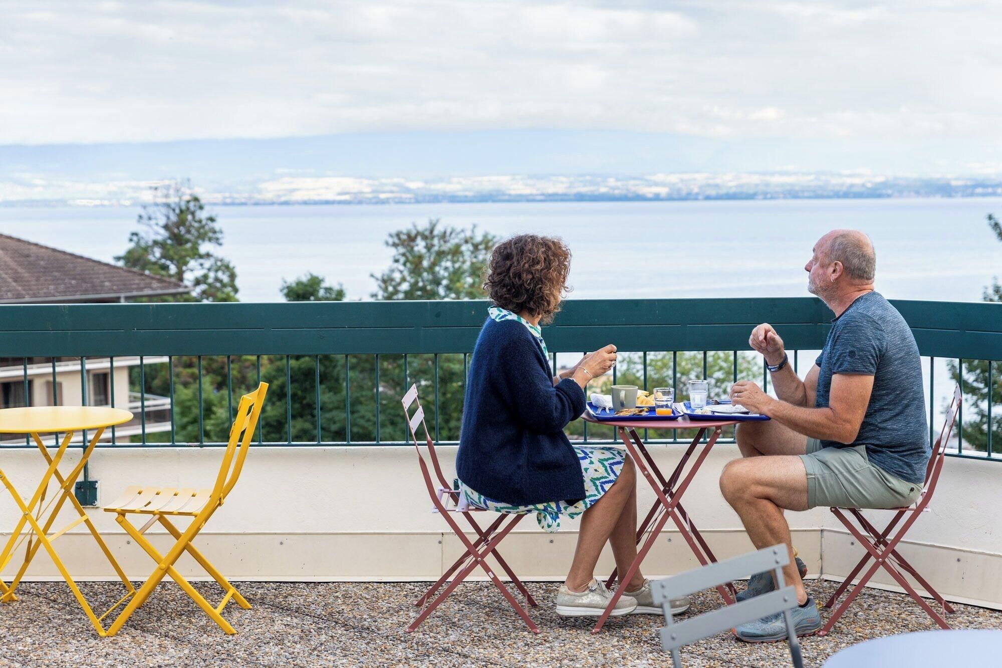Zwei Personen essen an einem Tisch im Freien mit Blick auf ein Gewässer und entferntes Land.