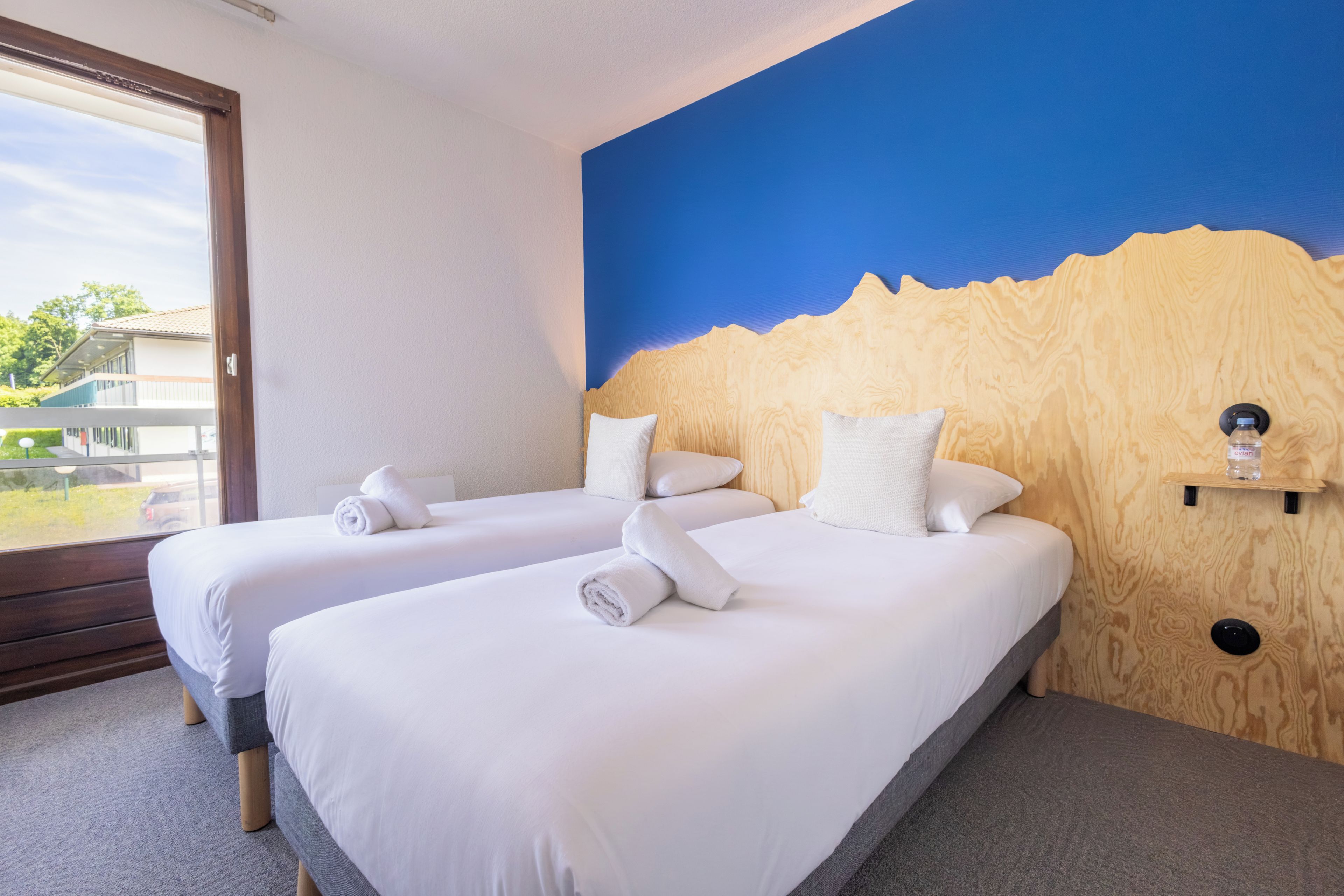 Habitación de hotel con dos camas, cabecera de madera, ventana con vistas y un pequeño estante con una botella.