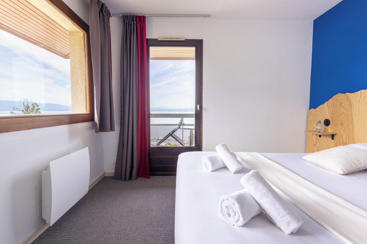 A hotel room with a bed, a radiator, a window with curtains, a view of the outdoors, and a wooden headboard.