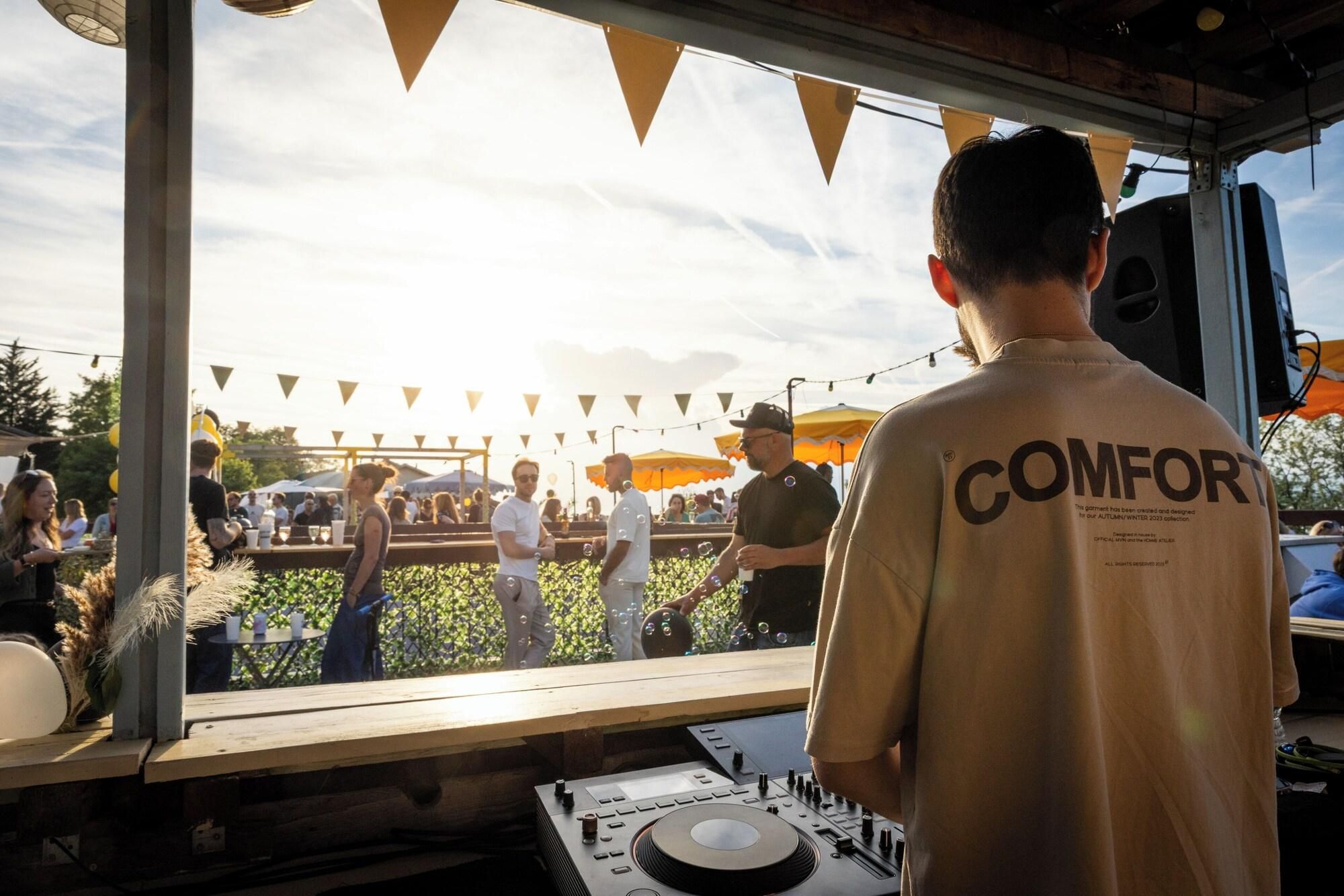A DJ booth with a mixer and turntables, a person wearing a 'COMFORT' branded shirt, and a crowd outside a venue with string lights and umbrellas.