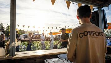 A DJ booth with a mixer and turntables, a person wearing a 'COMFORT' branded shirt, and a crowd outside a venue with string lights and umbrellas.