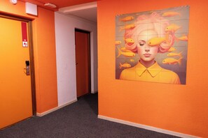 A hallway with orange walls, a door with a red emergency exit sign, and a wall mural with a person surrounded by fish. - Côté Lac Hostel (Neuvecelle)