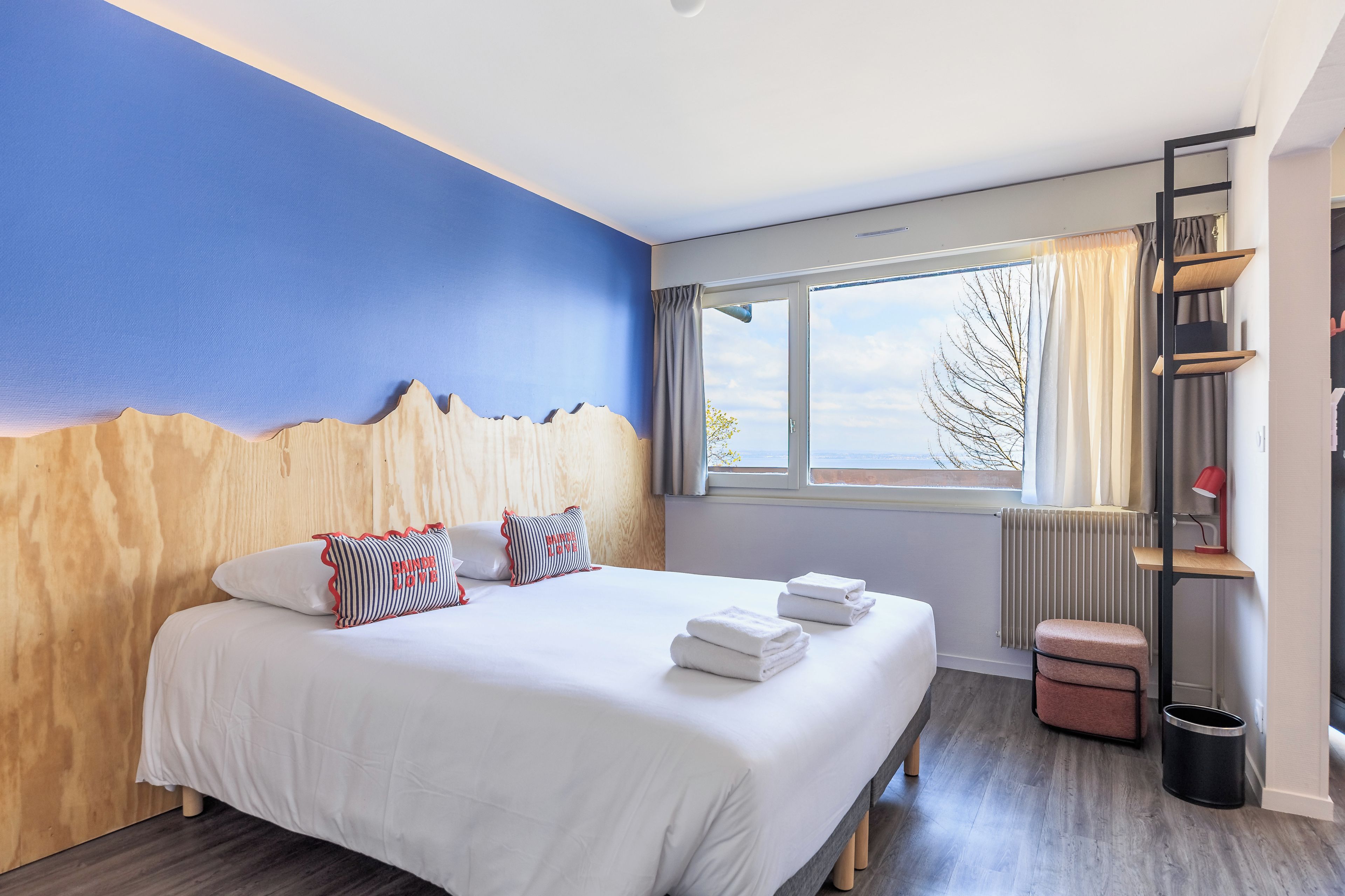 Quarto de hotel com cama, janela com cortinas, radiador, mesa pequena com abajur e lixeira.