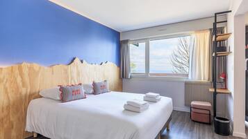 Quarto de hotel com cama, janela com cortinas, radiador, mesa pequena com abajur e lixeira.