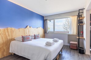 A hotel room with a bed, a window with curtains, a radiator, a small table with a lamp, and a trash can. - Côté Lac Hostel (Neuvecelle)