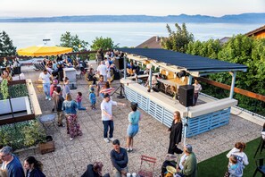 An outdoor patio with seating, a stage, and a view of the lake. - Côté Lac Hostel (Neuvecelle)