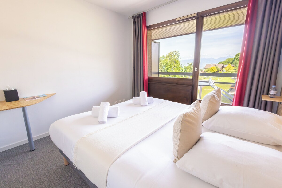 Ein Hotelzimmer mit einem Bett, zwei Kissen, einem Nachttisch mit Taschentuchbox und einem Fenster mit Vorhängen.