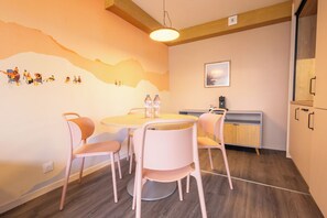 A dining area with a round table, four chairs, a wall mural, a kitchen area, and a hanging light fixture. - Côté Lac Hostel (Neuvecelle)