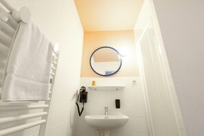 A narrow bathroom with a white sink, a towel rack, and a mirror with a blue frame. - Côté Lac Hostel (Neuvecelle)