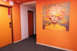 A hallway with orange walls, a door with a red emergency exit sign, and a wall mural with a person surrounded by fish. - Côté Lac Hostel (Neuvecelle)