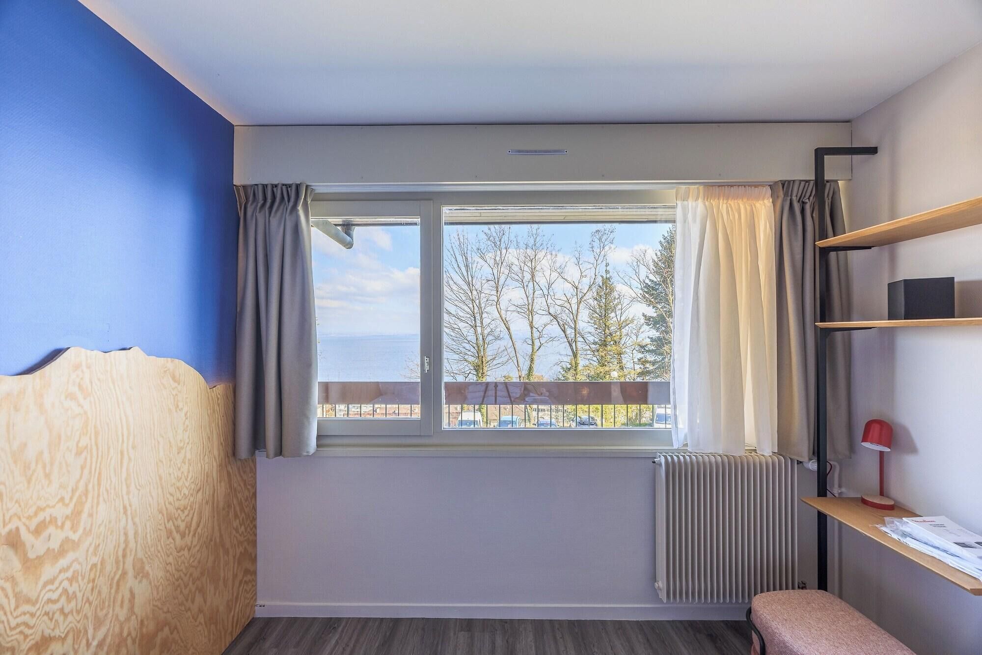 Ein Zimmer mit Fenster, Heizkörper, einem Holzkopfteil, einer Bank und einem Regal mit einer Lampe.