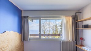 A room with a window, a radiator, a wooden headboard, a bench, and a shelf with a lamp. - Côté Lac Hostel (Neuvecelle)
