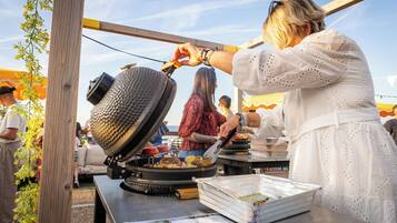 Una persona cocinando en una parrilla portátil, con una sartén y una olla, rodeada de una mesa con alimentos y un menú.