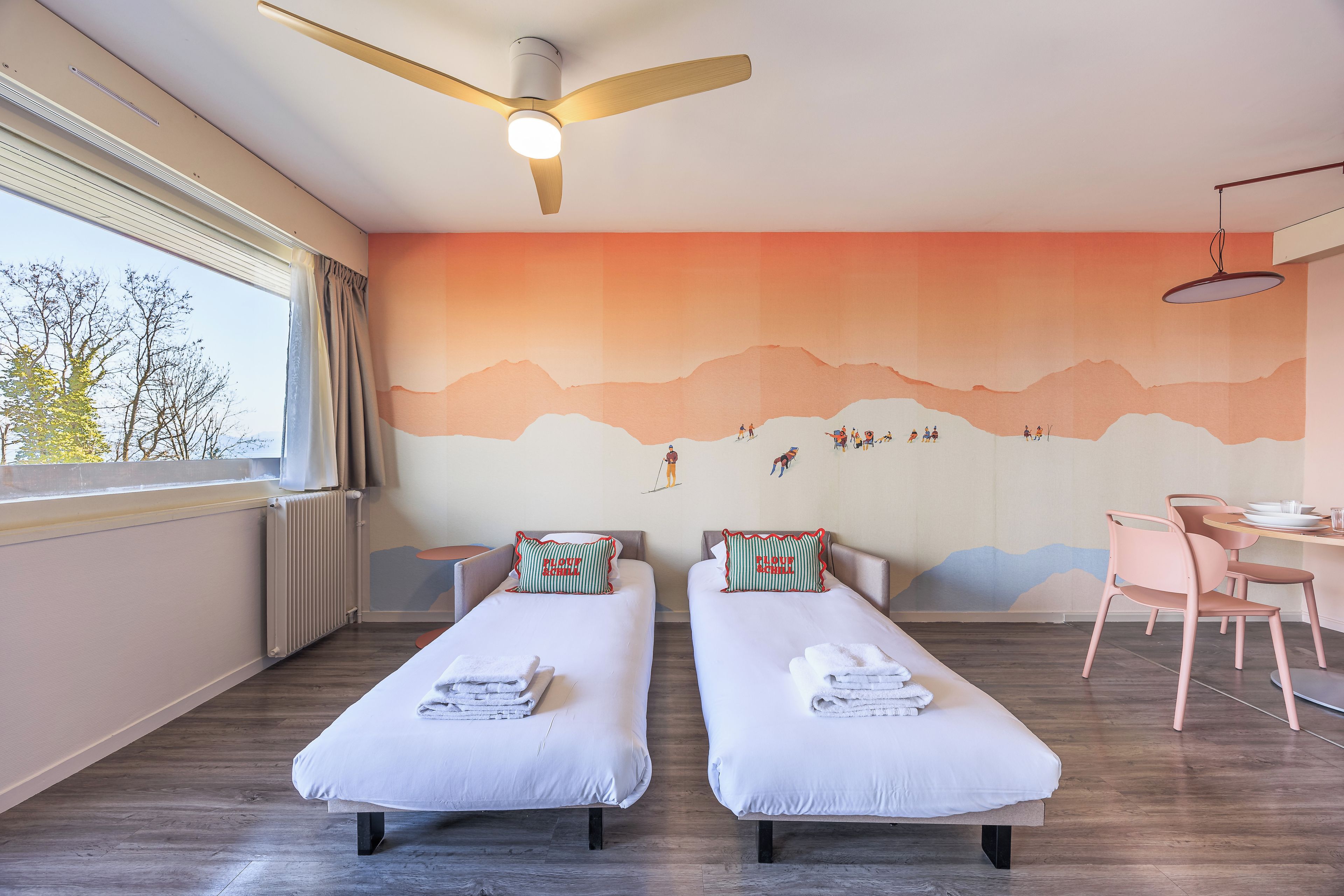 A hotel room with two single beds, a desk, a chair, a ceiling fan, and a large window.