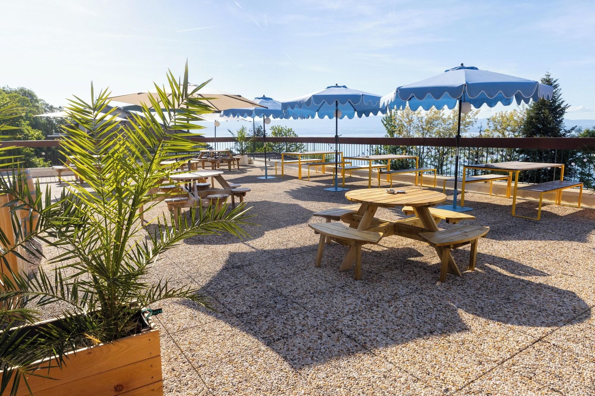 An outdoor patio with wooden tables and benches, surrounded by gravel, and blue umbrellas providing shade.