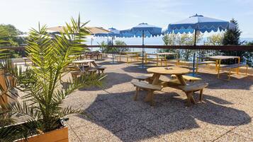 An outdoor patio with wooden tables and benches, surrounded by gravel, and blue umbrellas providing shade.