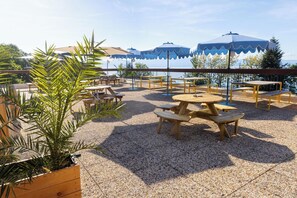 An outdoor patio with wooden tables and benches, surrounded by gravel, and blue umbrellas providing shade. - Côté Lac Hostel (Neuvecelle)