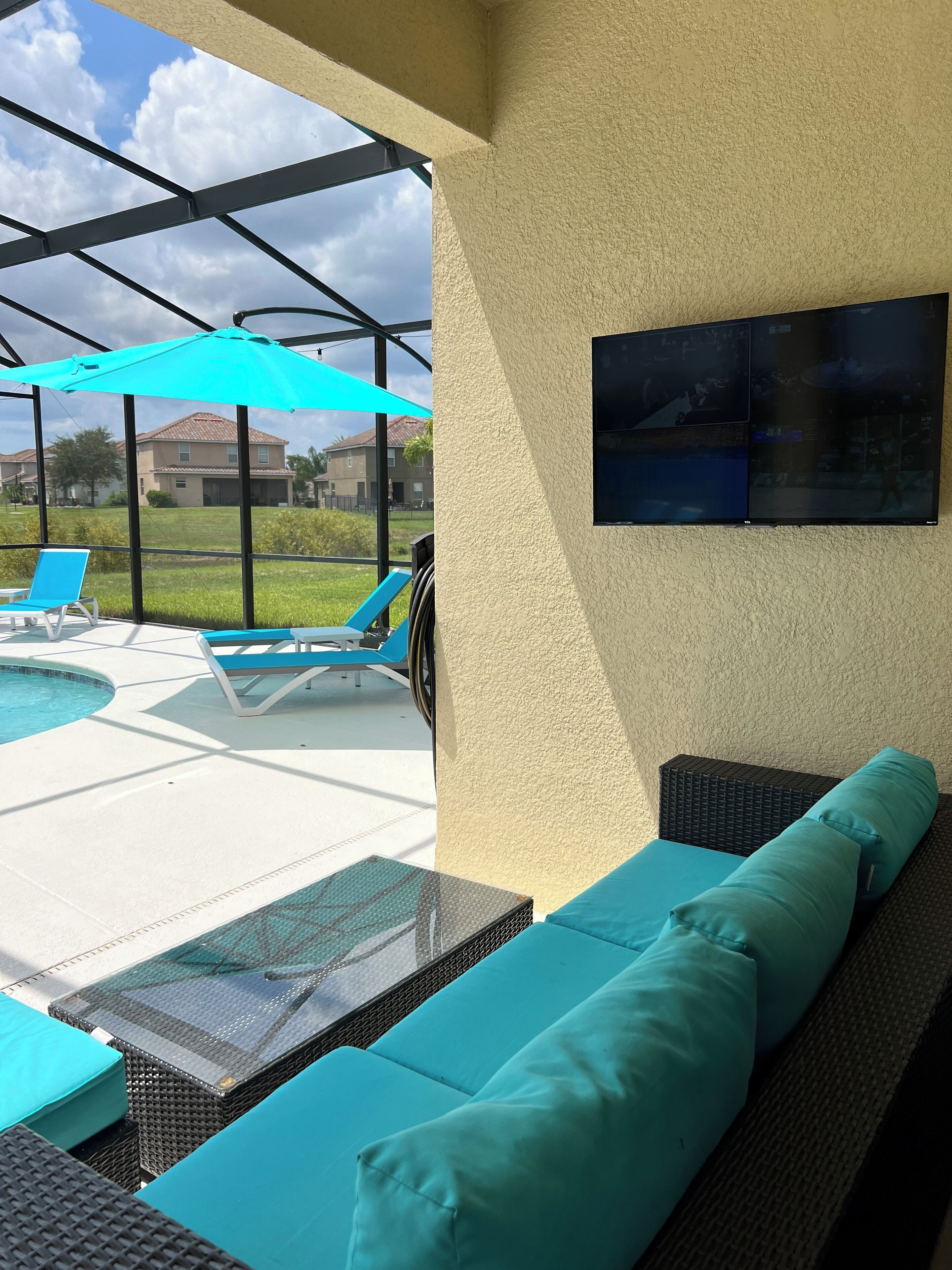 Un área junto a la piscina con un espacio de estar techado, un televisor montado en la pared y vistas del vecindario circundante.