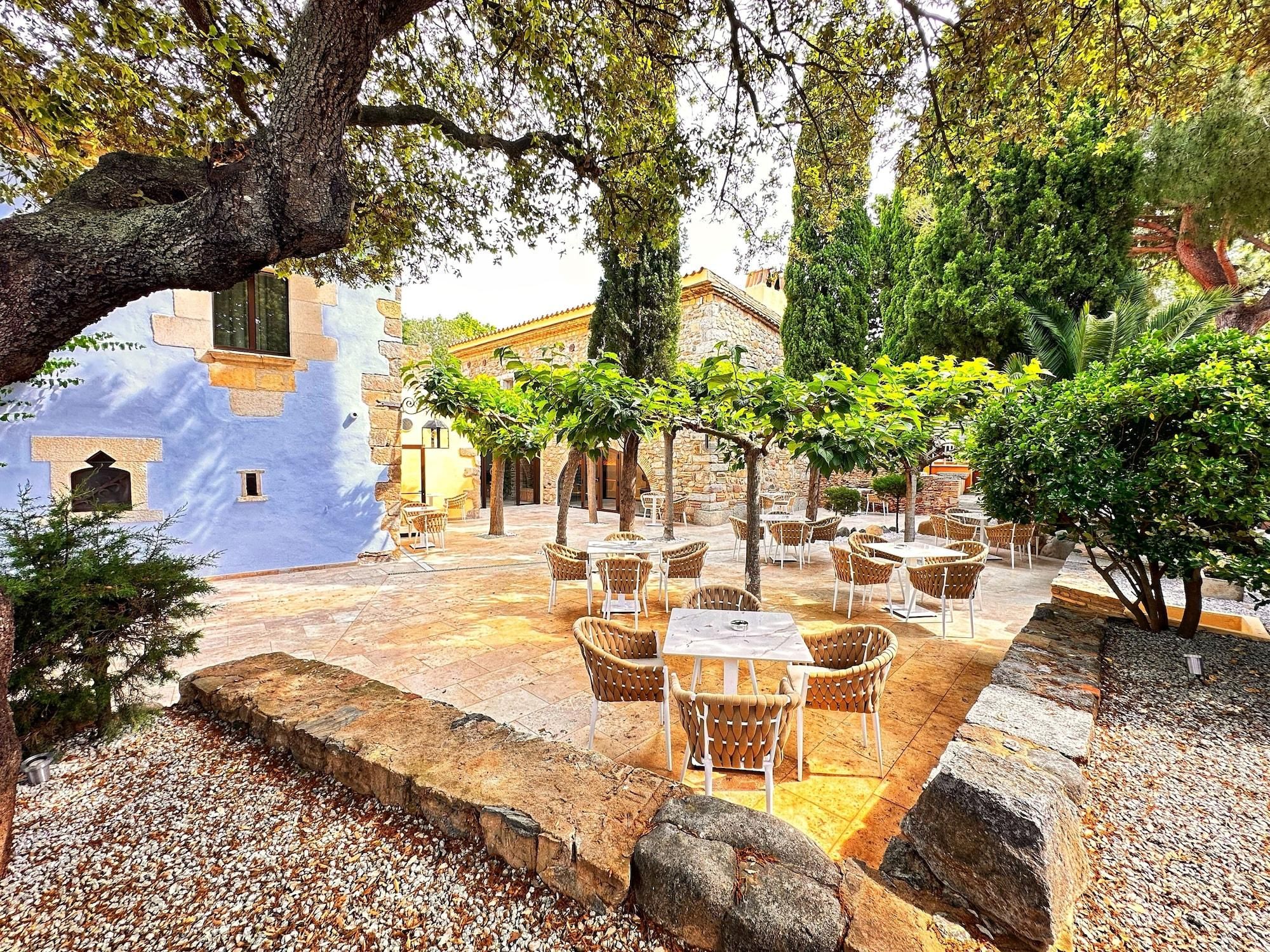 Un área de café al aire libre con mesas de piedra y sillas de mimbre, rodeada de árboles y un muro de piedra.