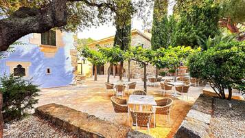 Un área de café al aire libre con mesas de piedra y sillas de mimbre, rodeada de árboles y un muro de piedra.