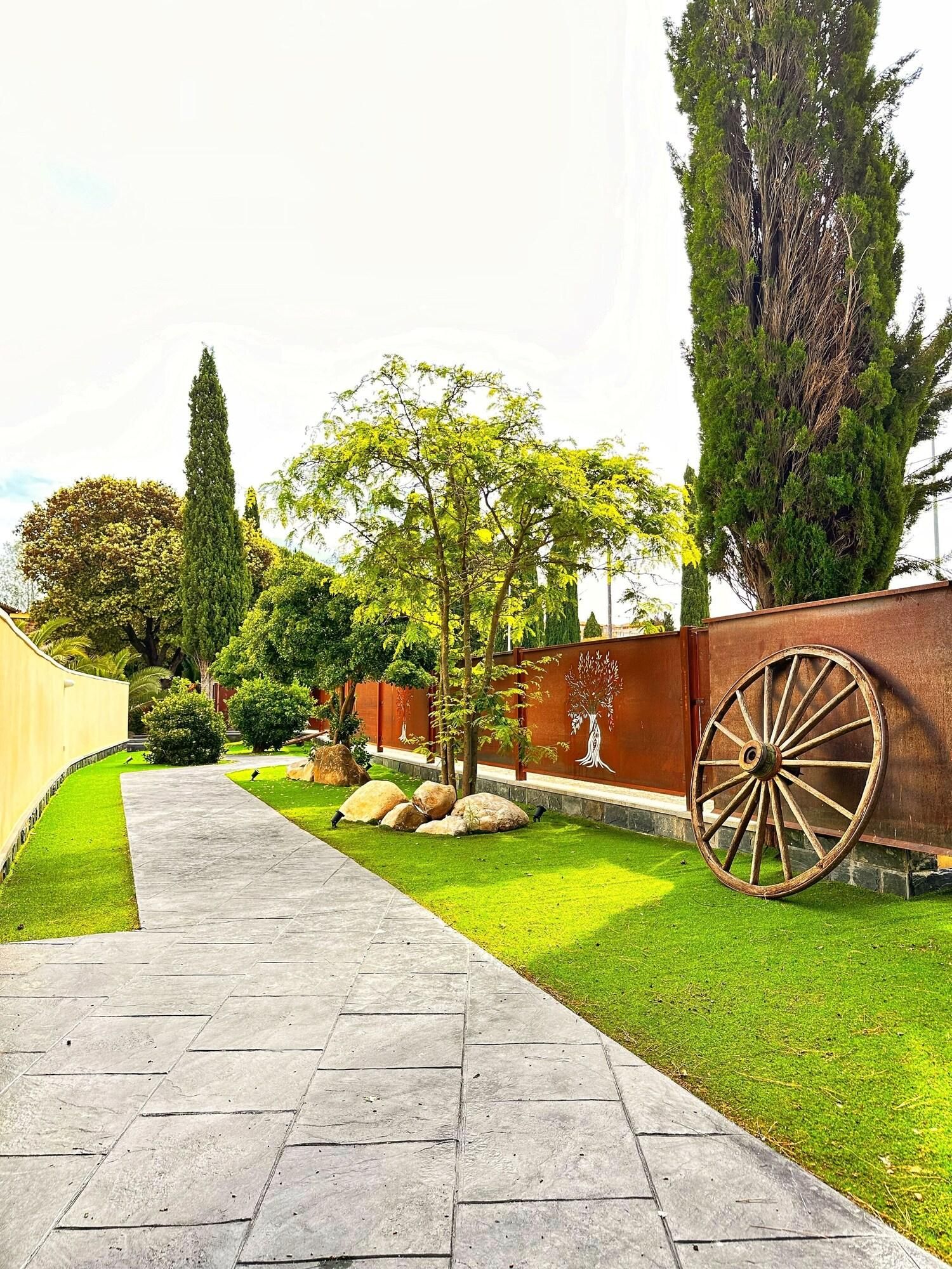 Um caminho pavimentado serpenteia por um jardim bem cuidado, com diversas árvores e arbustos.