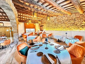 Un restaurante con paredes de piedra y vigas de madera, mesas dispuestas con platos y cubiertos, y sillas color naranja.